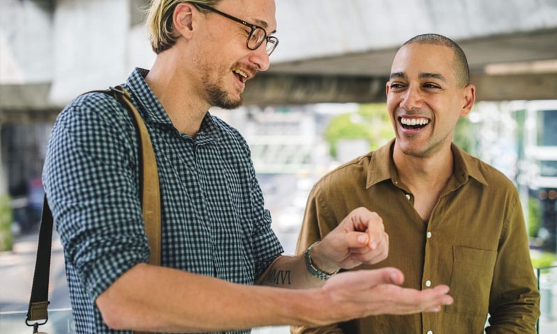 Two men laughing and talking outdoors under a bridge.