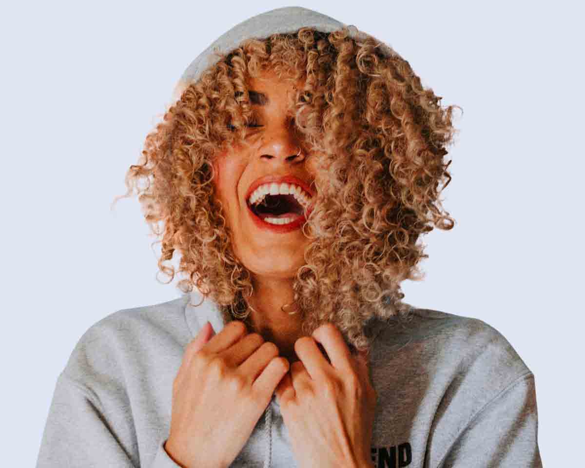 A joyful woman with curly hair laughing in a grey hoodie.