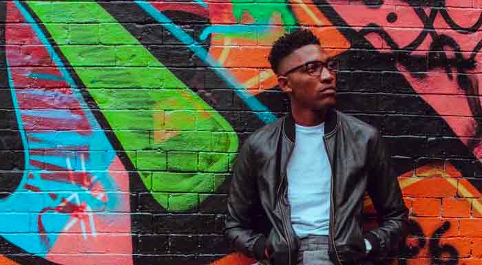 A man in glasses and leather jacket stands against a colorful graffiti wall.