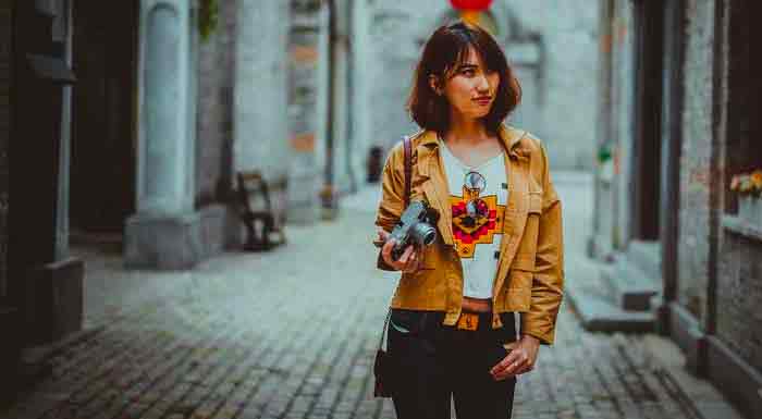 A young woman holding a camera in a cobblestone alley.