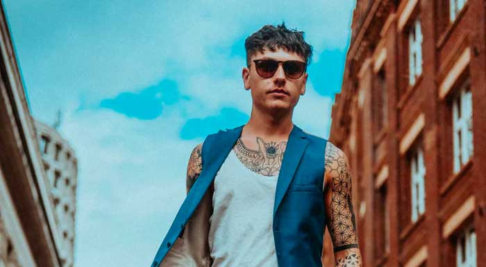 A tattooed man wearing sunglasses and a blue vest in an urban setting.