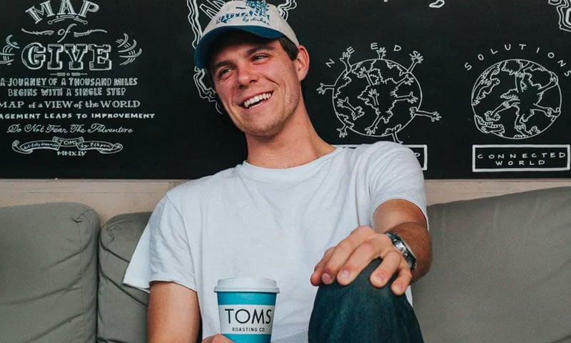 A smiling man in a white t-shirt and cap sitting on a couch holding a TOMS cup.